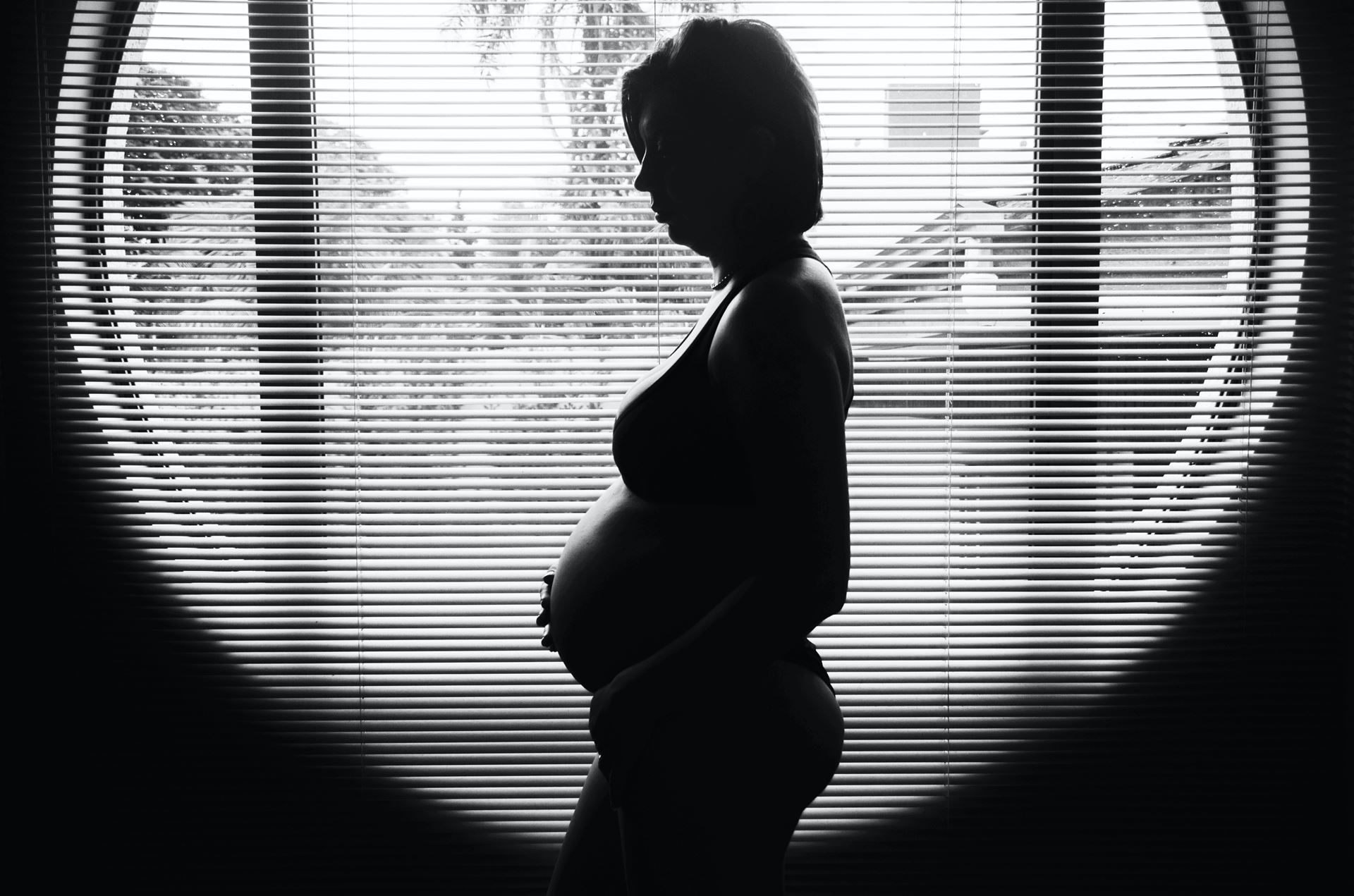 Black and white image of a pregnant individual's silhouette in front of a window with venetian blinds. Photo by Camila Cordeiro on Unsplash
